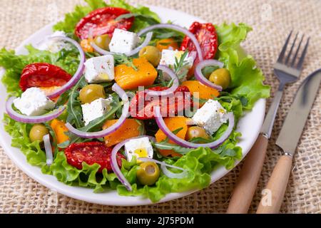 Warmer Salat mit Kürbis, Feta-Käse, getrockneten Tomaten, Oliven, Rucola und roter Zwiebel. Nahaufnahme. Stockfoto