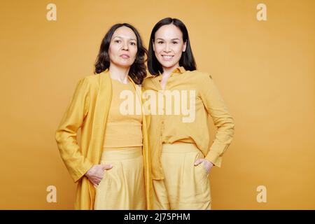 Porträt der erwachsenen kaukasischen Mutter und ihrer asiatischen Tochter in gelben Outfits mit Händen in Hosentaschen Stockfoto