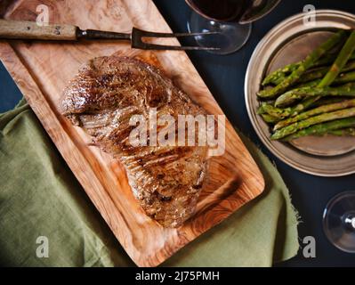 Ganze Knoblauchkraut-Sirloin auf einem Schneidebrett; Platte mit Spargel und Rotwein im Hintergrund Stockfoto