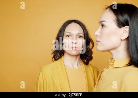 Reife kaukasische Mutter, die die Kamera anschaut, während ihre asiatische Erwachsene Tochter zur Seite schaut Stockfoto