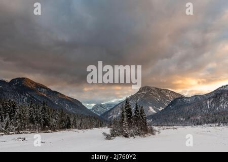 Das Risstal am Übergang zwischen Tirol und Deutschland im Karwendelgebirge mit dem ausgetrockneten Bachbett des Rissbaches und einer Insel mit Bäumen im Winter mit Schnee und einem bunten Sonnenuntergang mit dunklen Wolken. Stockfoto