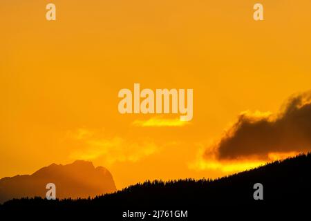 Sonnenuntergang auf Deutschlands höchstem Berg, der Zugspitze in Gold & Rot mit schwarzem Wald im Vordergrund. Eine Wolke schiebt sich von rechts ins Bild. Als Hintergrund vom Walchensee. Stockfoto