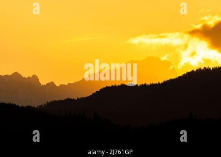 Sonnenuntergang auf Deutschlands höchstem Berg, der Zugspitze in Gold & Rot mit schwarzem Wald im Vordergrund. Eine Wolke schiebt sich von rechts ins Bild. Als Hintergrund vom Walchensee. Stockfoto