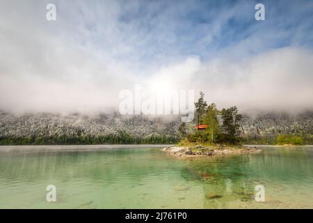 Eine kleine einsame Insel mit Blockhütte und rotem Dach, inmitten des schönen Eibsee unterhalb der Zugspitze in den bayerischen Alpen des Wettersteingebirges während der Eisheiligen - frischer Schnee im Hintergrund auf der Wald- und Wolkenatmosphäre. Stockfoto