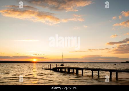 Sonnenuntergang mit Sonnenstrahlen auf einem hölzernen Steg am Ufer des Ammersee. Leichte Wellen plätschern das Wasser und im Hintergrund einzelne Segelboote. Stockfoto