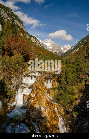 Der Karwendelbach bei Scharnitz auf dem Karwendelsteg im Herbst. Sonnenlicht lässt die Felsen leuchten, während im Hintergrund die schneebedeckten Gipfel vor einem blauen Himmel stehen. Stockfoto