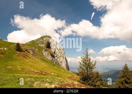 Ein Gleitschirm umkreist den Vorderer Kirchstein zwischen Brauneck und Benediktenwand im bayerischen Voralpenland, der kleine Gipfel steht vor einer hellen Wolke am sonst blauen Himmel, während der Athlet ganz nah daran fliegt. Stockfoto