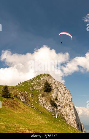 Ein Gleitschirm umkreist den Vorderer Kirchstein zwischen Brauneck und Benediktenwand im bayerischen Voralpenland, der kleine Gipfel steht vor einer hellen Wolke am sonst blauen Himmel, während der Athlet ganz nah daran fliegt. Stockfoto