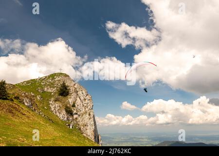 Ein Gleitschirm umkreist den Vorderer Kirchstein zwischen Brauneck und Benediktenwand im bayerischen Voralpenland, der kleine Gipfel steht vor einer hellen Wolke am sonst blauen Himmel, während der Athlet ganz nah daran fliegt. Stockfoto