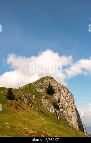 Ein Gleitschirm umkreist den Vorderer Kirchstein zwischen Brauneck und Benediktenwand im bayerischen Voralpenland, der kleine Gipfel steht vor einer hellen Wolke am sonst blauen Himmel, während der Athlet ganz nah daran fliegt. Stockfoto