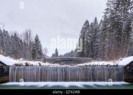 Ein Staudamm, im Hintergrund die typische Steinbrücke an der Obernach zwischen Wallgau und Walchensee in den bayerischen Alpen im Winter bei Schneefall und bewölktem Wetter Stockfoto