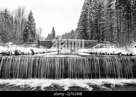 Ein Staudamm, im Hintergrund die typische Steinbrücke an der Obernach zwischen Wallgau und Walchensee in den bayerischen Alpen im Winter bei Schneefall und bewölktem Wetter Stockfoto