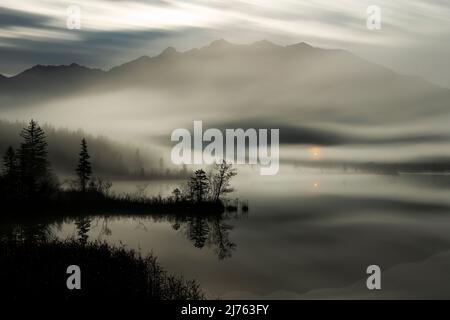 Die kleine Kapelle Maria rast bei Krün strahlt wie ein Hoffnungsschimmer durch den Nebel über dem kleinen Moorsee in den bayerischen Alpen. Im Hintergrund das Karwendelgebirge schattig, während die Landschaft durch den nicht sichtbaren Vollmond beleuchtet wird, im Vordergrund ein kleines Vorgebirge im Waser, mit Bäumen und Sträuchern. Stockfoto