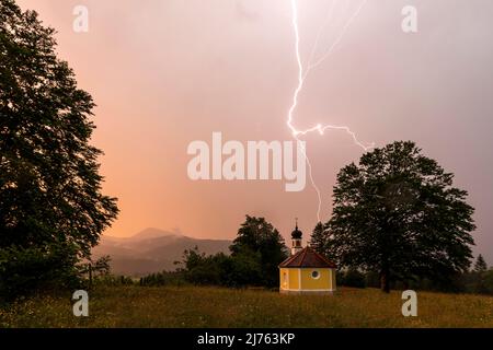 Gewitter mit Blitzen in der kleinen Kapelle Maria Rast im Werdenfelser Land in den bayerischen Alpen zwischen Mittenwald und Garmisch Partenkirchen, bei rötlich-abendlicher Atmosphäre. Stockfoto