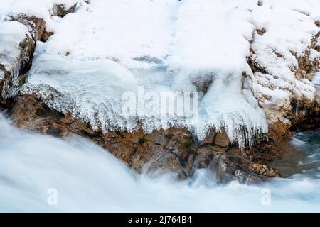 Schnelles Wasser des Rissbaches bildet Spray und Strom. Eisstrukturen und Felsen, die am Ufer des Gebirgsbaches gebildet werden, Grenzen das Bild ein. Aufgenommen im Winter in Tirol, Österreich im eng, nahe dem Ahornboden. Stockfoto