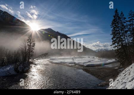 Schneelandschaft in den Isarauen, mit dem Fluss und einzelnen Fichten im Winter bei Sonnenuntergang an der Grenze zum Karwendel bei Vorderriss und Wallgau auf der Mautstraße. Der farbenfrohe Himmel nach Sonnenuntergang spiegelt sich teilweise im Wasser der Isar wider. Stockfoto