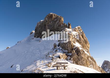 Die westliche Karwendelspitze im Karwendel oberhalb von Mittenwald und die Bergstation der Karwendelbahn, auf dem Passamani-Rundweg mit einer Bank im Winter in Schnee und Eis, unter blauem Himmel. Stockfoto