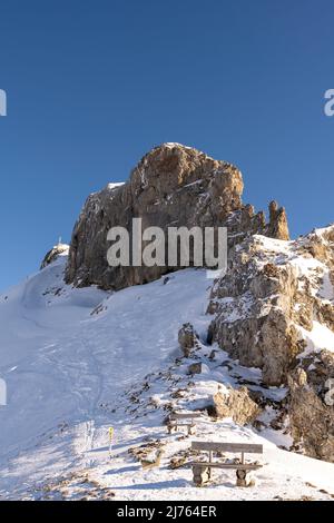 Die westliche Karwendelspitze im Karwendel oberhalb von Mittenwald und die Bergstation der Karwendelbahn, auf dem Passamani-Rundweg mit einer Bank im Winter in Schnee und Eis, unter blauem Himmel. Stockfoto