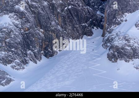 Im Schatten des östlichen Karwendelgipfels, im Rhonetal bei Hinterriss, Karwendel, führt der Zickzack hinauf zu den Felswänden, kleine Skifahrer nutzen die Pisten für schattige, wilde Abfahrten, während die kalten Felswände über ihnen ragen. Stockfoto