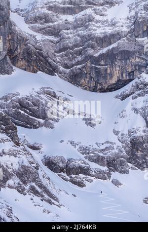 Im Schatten des östlichen Karwendelgipfels, im Rhonetal bei Hinterriss, Karwendel, führt der Zickzack hinauf zu den Felswänden, kleine Skifahrer nutzen die Pisten für schattige, wilde Abfahrten, während die kalten Felswände über ihnen ragen. Stockfoto