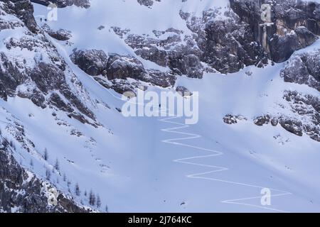 Im Schatten des östlichen Karwendelgipfels, im Rhonetal bei Hinterriss, Karwendel, führt der Zickzack hinauf zu den Felswänden, kleine Skifahrer nutzen die Pisten für schattige, wilde Abfahrten, während die kalten Felswände über ihnen ragen. Stockfoto