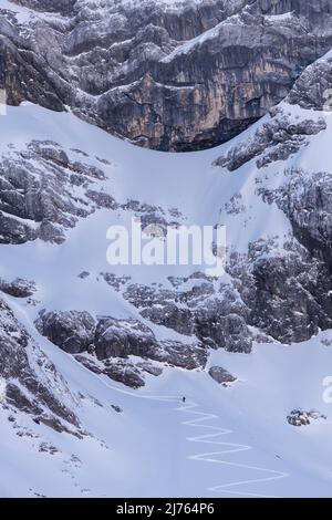 Im Schatten des östlichen Karwendelgipfels, im Rhonetal bei Hinterriss, Karwendel, führt der Zickzack hinauf zu den Felswänden, kleine Skifahrer nutzen die Pisten für schattige, wilde Abfahrten, während die kalten Felswände über ihnen ragen. Stockfoto