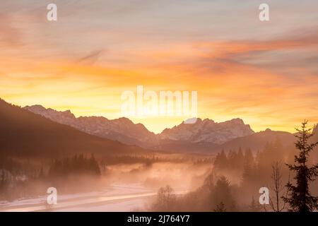 Die Isar windet sich durch die mit Reif bedeckte und im Winter teilweise verschneite Landschaft. Im Hintergrund glühen die Zugspitze und ein fantastischer Abend über den Wettersteingebirge. Stockfoto