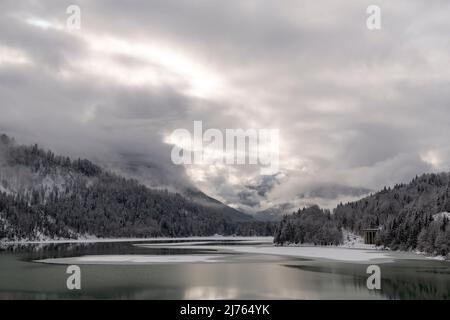 Der Sylvensteinspeichersee am Rande des Karwendelgebirges in den bayerischen Alpen im Winter mit Schnee und teilweise mit Eis, sowie Wolkenatmosphäre. Stockfoto