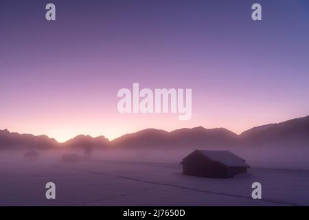 Neblige Atmosphäre zwischen Krün und Wallgau im Werdenfelser Land im Winter. Dichter Nebel liegt über den verschneiten Wiesen mit Stadln, im Hintergrund die Wettersteingebirge und die Zugspitze im violetten Abendrot mit wolkenlosem Himmel. Stockfoto