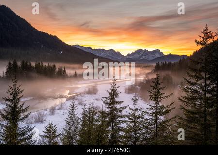 Die Isar windet sich durch die mit Reif bedeckte und im Winter teilweise verschneite Landschaft. Im Hintergrund glühen die Zugspitze und ein fantastischer Abend über den Wettersteingebirge. Stockfoto