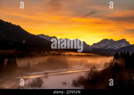 Die Isar windet sich durch die mit Reif bedeckte und im Winter teilweise verschneite Landschaft. Im Hintergrund glühen die Zugspitze und ein fantastischer Abend über den Wettersteingebirge. Stockfoto