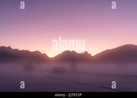 Neblige Atmosphäre zwischen Krün und Wallgau im Werdenfelser Land im Winter. Dichter Nebel liegt über den verschneiten Wiesen mit Stadln, im Hintergrund die Wettersteingebirge und die Zugspitze im violetten Abendrot mit wolkenlosem Himmel. Stockfoto