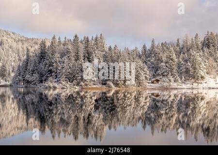 Die kleine Hütte gegenüber der Wasserwacht am Lautersee bei Schnee und leichten Wolken an einem sonnigen Wintertag. Das Seeufer spiegelte sich im Wasser des kleinen Bergsees wider Stockfoto
