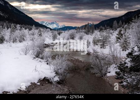 Ein kleiner Nebenstrom der Isar windet sich im Winter durch die verschneite Landschaft, die von Reif bedeckt ist. Im Hintergrund glühen die Zugspitze und ein fantastischer Abend über den Wettersteingebirge. Stockfoto