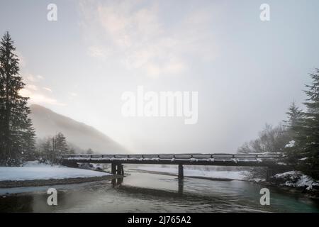 Die kleine Brücke bei Vorderriss neben der Mautstraße im Schnee bei Dämmerung im dichten Nebel. Die Isar fließt zum Betrachter hin. Stockfoto