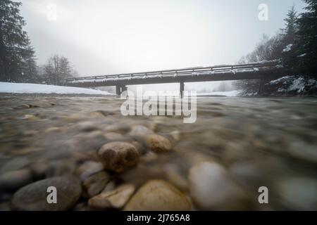 Die kleine Brücke bei Vorderriss neben der Mautstraße im Schnee bei Dämmerung im dichten Nebel. Die Isar fließt zum Betrachter hin. Stockfoto