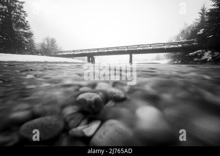 Die kleine Brücke bei Vorderriss neben der Mautstraße im Schnee bei Dämmerung im dichten Nebel. Die Isar fließt zum Betrachter hin. Stockfoto