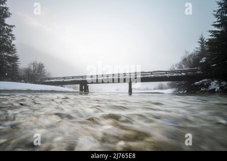 Die kleine Brücke bei Vorderriss neben der Mautstraße im Schnee bei Dämmerung im dichten Nebel. Die Isar fließt zum Betrachter hin. Stockfoto