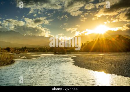 Auf dem Flussbett der Isar bei Wallgau im Werdenfelser Land in Bayern bei Wallgau bildet sich bei Sonnenuntergang ein Sonnenstern. Im Hintergrund das Wettersteingebirge mit der Zugspitze, mit schönem Himmel und goldenem Licht im Herbst. Stockfoto