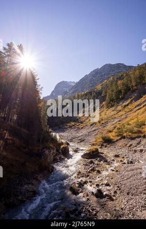 Sonne mit Sonnenstrahlen am herbstlichen Johannisbach im gleichnamigen Tal im Karwendel bei Hinterriss, Österreich / Tirol. Der Bach schlängelt sich um eine Biegung, die gut die Bodenerosion zeigt... Stockfoto