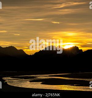 Sonnenstern der untergehenden Sonne auf der Zugspitze. Im Vordergrund die leuchtende Isar am sogenannten Isarsteg bei Wallgau im Werdenfelser Land. Stockfoto