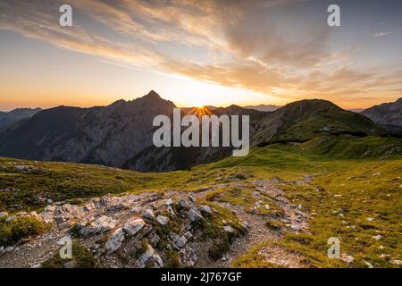 Wanderweg mit Wandermarkierungen, im Hintergrund die Mondscheinspitze mit einem tollen Sonnenstern mit hellen Wolken am sonst blauen Himmel und goldenen, grünen Bergwiesen. Der Weg führt über einen Grat zur Plumsjochhütte von Kompar im Herzen des Karwendels. Stockfoto