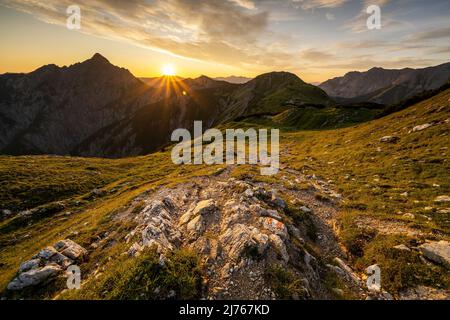 Wanderweg mit Wandermarkierungen, im Hintergrund die Mondscheinspitze mit einem tollen Sonnenstern mit hellen Wolken am sonst blauen Himmel und goldenen, grünen Bergwiesen. Der Weg führt über einen Grat zur Plumsjochhütte von Kompar im Herzen des Karwendels. Stockfoto