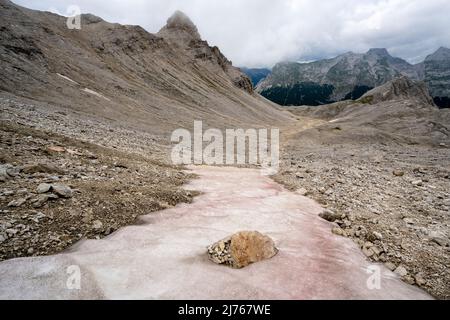 Im Hochgebirge der alpinen Hinterau-Vomper-Gebirgskette in Karwendel bleibt Staub und Sand der roten Sahara auf dem Schnee. Im Hintergrund die unwirtliche Landschaft und dichte Wolken. Stockfoto