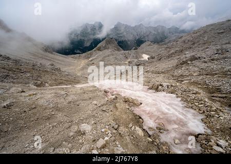 Im Hochgebirge der alpinen Hinterau-Vomper-Gebirgskette in Karwendel bleibt Staub und Sand der roten Sahara auf dem Schnee. Im Hintergrund die unwirtliche Landschaft und dichte Wolken. Stockfoto