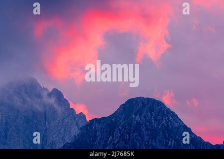 Spektakulär leuchtende Wolken bei Sonnenuntergang auf der östlichen Karwendelspitze, der genau hinschaut, entdeckt das Gipfelkreuz. Aufgenommen auf der Abfahrt von der Birkkarspitze in Karwendel, Tirol. Stockfoto