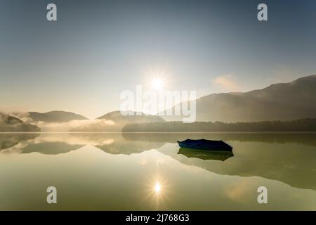 Ein einziges Ruderboot mit Plane ist am Sylvensteinspeicher vor Anker, während die Sonne am frühen Morgen mit leichtem Nebel ihre Strahlen über den See sendet, die sich im Wasser spiegeln. Stockfoto