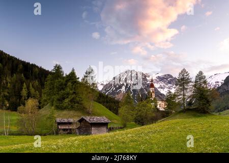 Alte Holzmühlen in Obernberg am Brenner, die historische Nikolaikirche im Hintergrund und Blumenwiesen im Vordergrund. Stockfoto