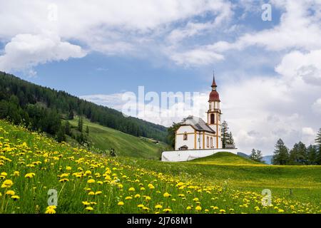 Die historische Nikolaikirche in Obernberg am Brenner in Tirol. Im Vordergrund eine Blumenwiese mit Delaionblüten, im Hintergrund blauer Himmel und Wolken. Stockfoto