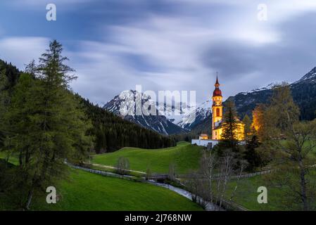 Alte Holzmühlen in Obernberg am Brenner, die historische Nikolaikirche im Hintergrund und Blumenwiesen im Vordergrund. Stockfoto